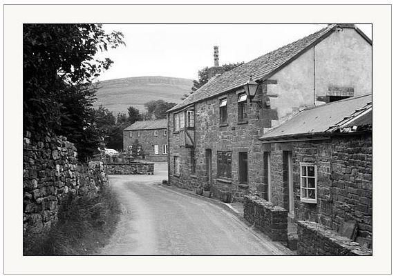 Black and white photograph showing the view down an English country lane with old cottages to the right and a hill in the distance.