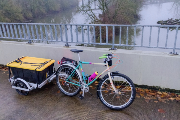 A colorful upright bike towing a cargo trailer with a large storage bin on a multi-use path crossing the Columbia Slough on a rainy day. 