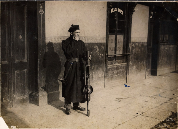 A Black and Tan in Ireland, casually holding a Lewis gun and smoking a cigarette in front of a drinking establishment. Dressed in a military suprlus great coat, a black tam o'shanter a thick leather belt and low slung holster, with regular civies underneath.