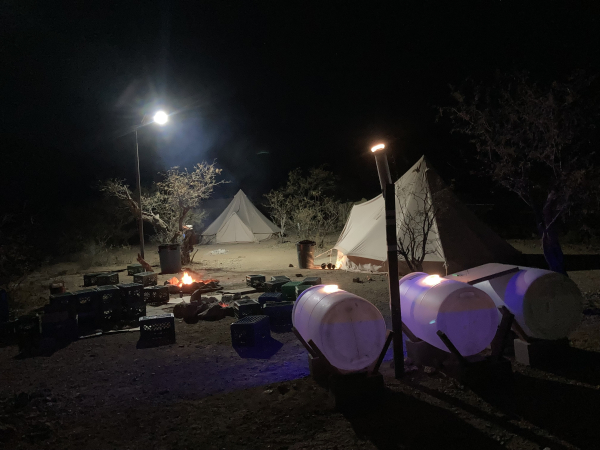 A nighttime scene of a camp. There is a burning campfire, a small solar powered street light up on a pole and two white canvas semi-conical tents. In the foreground are three large white plastic water barrels sitting in cradles on their side. There are no people in the camp at present. There are plastic milk crates used for seating scattered about, along with pieces of old carpeting and cardboard for people to sit on.