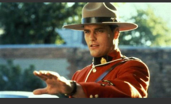 The actor Paul Gross dressed as a Mountie in character as Benton Fraser from the TV show due south. He’s a white man wearing a brown hat, a red dress uniform and holding out his hand as if to de-escalate a tension out of shot. The background is a wall and trees.