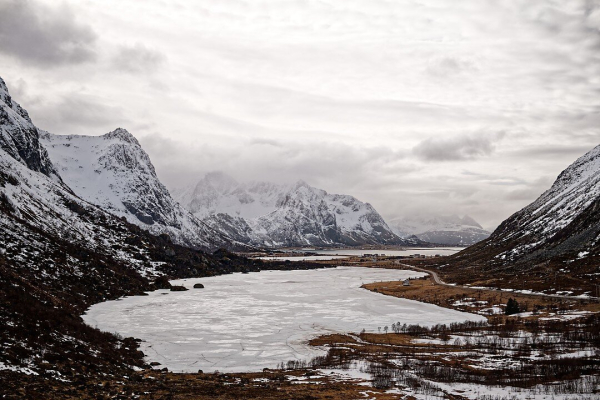 The image shows a winter valley with a partially frozen lake surrounded by snow-covered mountains under an overcast sky. A winding road follows the lake's edge, leading to a distant settlement, while jagged peaks and low clouds create a moody, serene atmosphere.