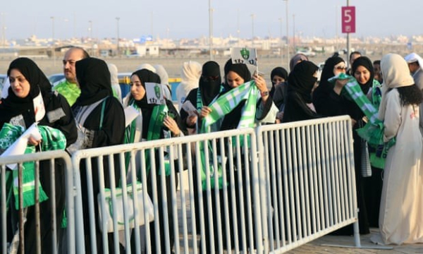 Arabic women in a mix of niqabs and hijabs queuing to enter the stadium whilst putting on green and white football scarves.