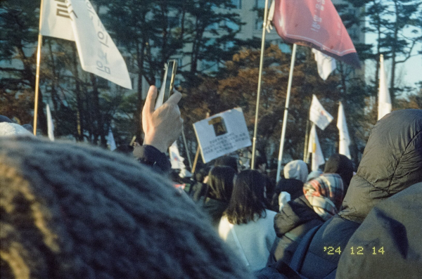 시위 군중과 무수히 많은 깃발 사이로 이 광경을 찍기 위해 스마트폰을 들고 있는 손이 보인다. 2024년 12월 14일.