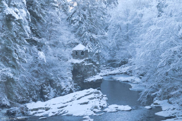 A snowy river scene, with a hexagonal stone folly building on a large rock, overlooking the water.