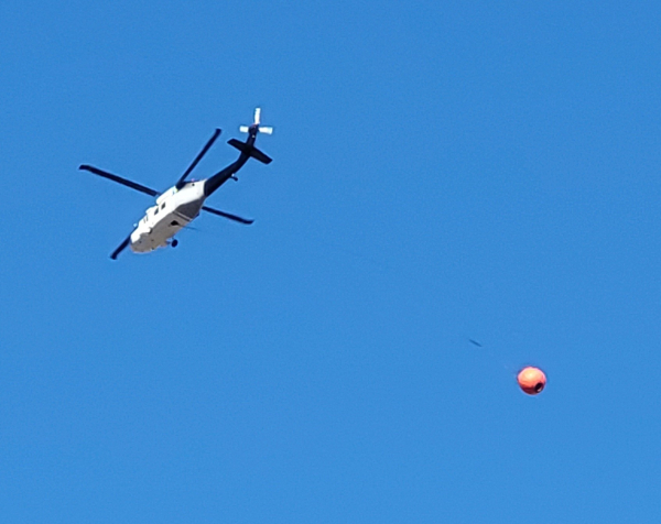 White and black helicopter with big orange bucket dragging on a long line behind