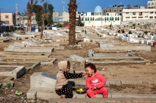 A child feeds another a spoonful of food as they sit on top of graves at a cemetery where families displaced by conflict are taking shelter, in Deir el-Balah in the central Gaza Strip on January 12, 2025 [Eyad Baba/AFP]