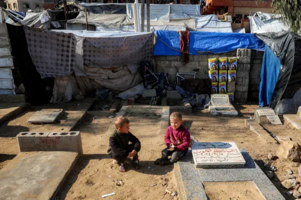 [Eyad Baba/AFP] 

Two children sit in the sand between graves.