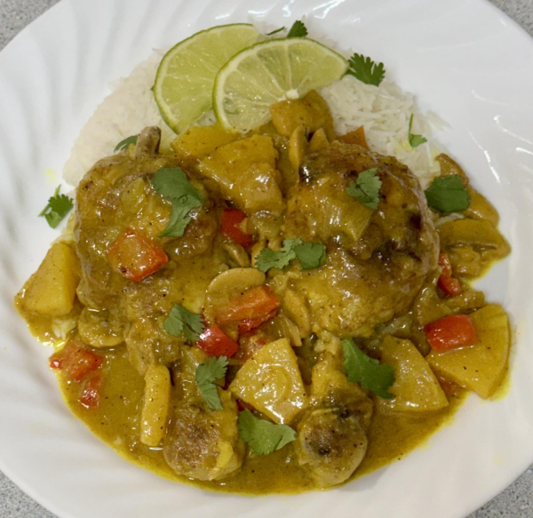 A white plate with two chicken legs covered with yellow curry sauce,pineapple chucks,red pepper,mushrooms,cilantro. 