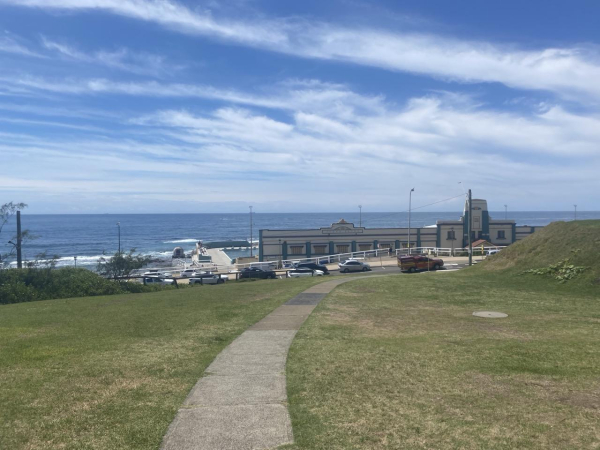 Looking towards the front of the Ocean Baths and out to sea from a grassy hill above. Over half of the heritage art deco facade of the Baths is visible, cream with turquoise and rose highlights. The turquoise concrete bleachers and part of the lap pool are behind on the left. Sky is blue streaked with white clouds on the diagonal.