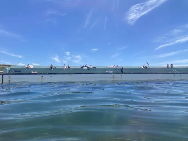 View of the turquoise concrete bleachers from in the main pool of the ocean baths. Green blue water, white pool wall, turquoise bleachers that bathers are sitting or lying on, blue sky with only wispy streaks of white cloud.