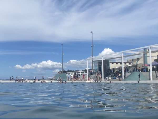 Taken looking southwesterly towards the Ocean Baths buildings while swimming in the main pool. 

Blue water with shiny reflections and over 20 swimmers along the white pool edge. More people sitting under shade shelters or walking along the concourse.

There is a thin sheet of white cloud above the buildings, blue sky then puffier white clouds off behind the baths complex. 