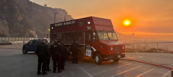 Against a fiery red sky and smoke-hazed sun, public servants dressed in black line up for a free hot meal from a red food truck in a parking lot on the Pacific Ocean in Los Angeles County. Photo credit: WCK