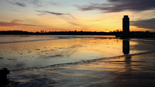 Strand des Strandbades in den Königswiesen an der Schlei. Das Hochwasser der letzten Tage liegt wie ein Tuch aus Eis über dem Sandstrand. Das Wasser ist spiegelglatt zugefroren. Auf dem Eis strahlen die Farben des Sonnenuntergangs in rot und gelb und blau. Im Hintergrund der Wikingturm, schwarz und dunkel, so auch sein Spiegelbild.