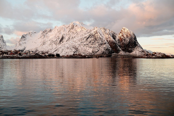 This image captures a snow-covered mountain range with warm sunlight reflecting off its peaks, creating a soft golden glow. The tranquil water in the foreground mirrors the sky and mountains, enhancing the serenity of the scene.