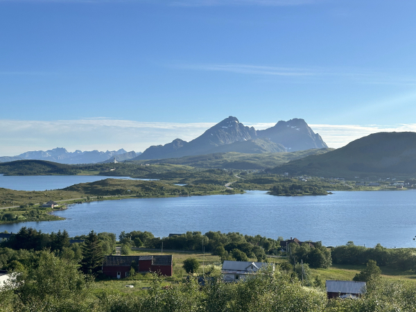 A panoramic landscape view in Lofoten, with pointed sharp mountains in the distance, remains of snow flecks just visible, behind two blue lakes and the green of midsummer