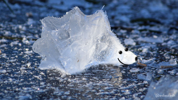 A photo of the surface of a frozen pond with a big chunk of ice in the centre. The ice has been warped slightly so the left hand side is slightly spiky, and the right goes to a point. Eyes, mouth and nose have been drawn on the point so it resembles some kind of hedgehog shaped ice creature. It is quite cute. Cold, but cute.