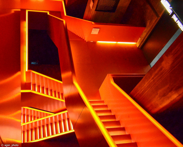 Blick hinunter in ein mehrstöckiges Treppenhaus ohne Fenster, die Treppen sind ganz rot, die Handläufe und Stufen haben gelbe Leuchtstreifen. /
View down into a multi-story staircase without windows, the stairs are all red, the handrails and steps have yellow luminous strips.
