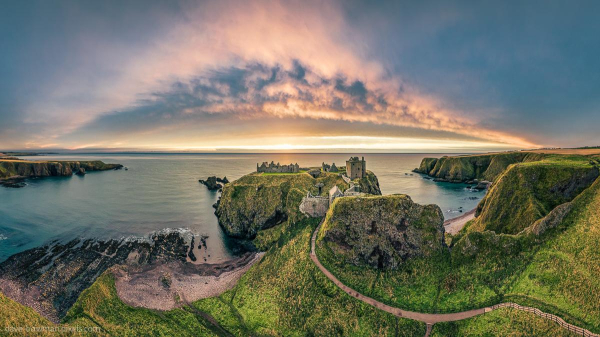 A photograph of Dunnottar Castle at sunrise, captured from above, highlighting its striking position on the rugged Aberdeenshire coastline. The warm light of dawn bathes the castle and surrounding cliffs, while soft clouds arch across the sky, enhancing the scene’s grandeur. Below, a winding path leads to the castle, inviting a closer look at this remarkable blend of natural beauty and historic architecture.