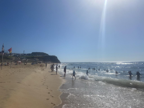 Looking northerly along where the water meets the sand at Bar Beach. Surf lifesaving flags flying red and yellow: beach open. Fair number of folks frolicking in or on the edges of the water. Calm day and a rising tide. Blue sky, darker blue ocean and sun glittering everywhere there's water. 