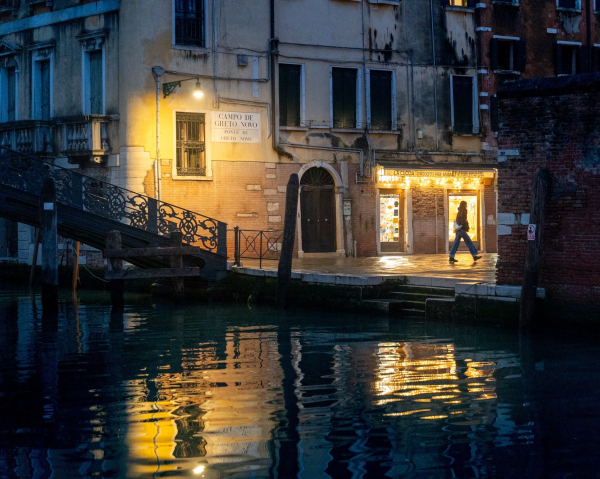 A person walks along a wet cobblestone street in Venice, illuminated by soft yellow lights. A shop displays colorful items, and a nearby bridge leads into the scene. The water reflects the lighting and surroundings, creating a serene evening atmosphere.