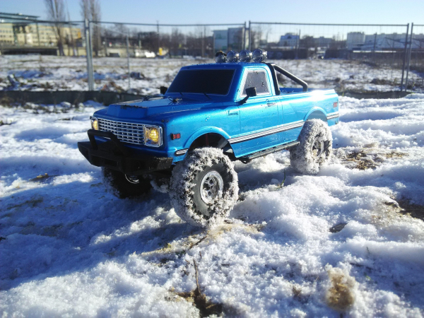 Das blaue ferngesteuerte Auto steht auf einer schneebedeckten Wiese. 
Die Reifen sind mit Schneeflocken eingedeckt. 
Das Fahrzeug steht mit der Front nach links, von rechts scheint die Wintersonne. 