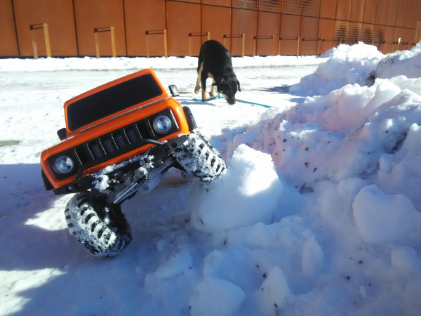 Das große orangefarbene ferngesteuerte Auto (RedCat International Scout 2 Gen8) steht mit einem Reifen auf einem großen, festen Schneeballen. 
Die Achse ist dabei stark verschränkt. 
Von rechts vorne scheint die Sonne. 
Im Hintergrund schnüffelt Tim auf dem Boden. 