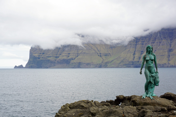 Bronze statue of a selkie/woman in front of a seascape with cliffs in the distance: “Kópakonan” selkie-statue, Mikladalur, Kalsoy, Faroe Islands (© kallerna, via WikiCommons, CC BY-SA 4.0)