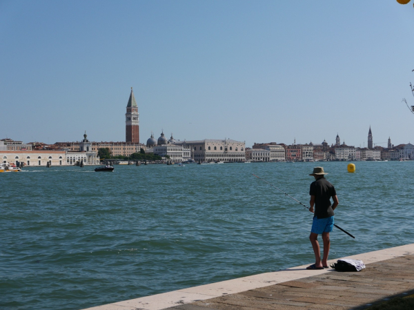 A fisherman wearing shorts and flip-flops faces away from the camera. Across a stretch of open  water can be seen some famous Venice landmarks, including the Doge's Palace.