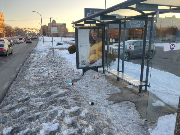 A bus station, covered in filthy snow and trash, next to a completely clear road. It's a stop for the #70 on Grand in St. Louis, Missouri.