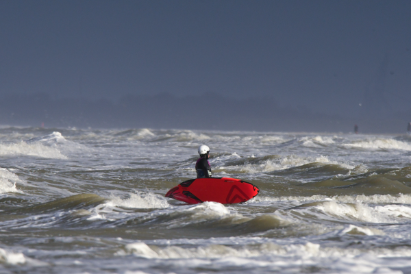 Surfer wearing a red windsurf board on the side of her body in the cold surf in winter. Black neoprene suit and white helmet protect her. The sea is churning.