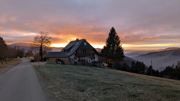 A two-story house with a gabled roof and dormer windows sits on a grassy hill overlooking a valley at sunset.  The sky is a mix of orange, pink, and purple hues. A tall evergreen tree stands to the right of the house. A paved road leads away from the house towards the left, and a bare deciduous tree is visible to the left of the house. Distant hills and mountains are visible in the background under the colorful sky.