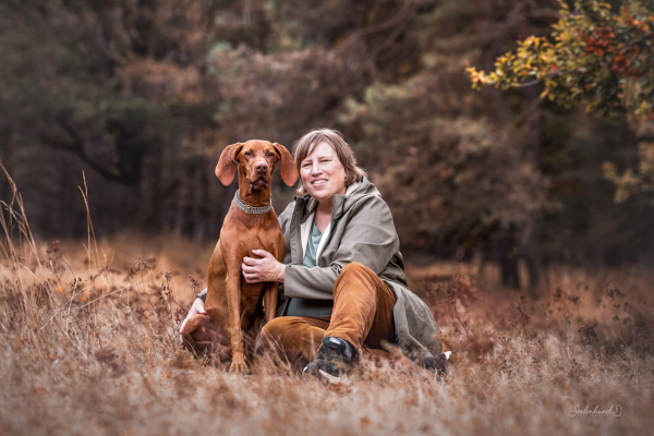 Eine Frau sitzt neben einem rotbraunen Hund im Gras, dahinter ist Wald zu sehen.