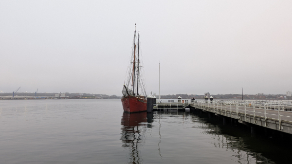 Ein Traditions-Segelschiff mit rotem Rumpf liegt an der Reventlou-Brücke / Kieler Förde. 
Es ist ein grauer, nebliger Tag, das Wasser ist ruhig. 