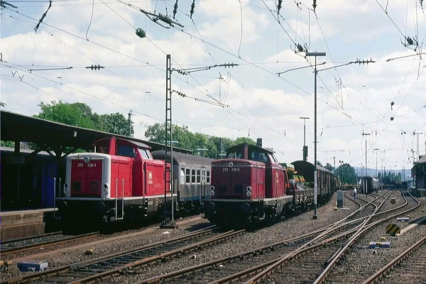 Das Bild zeigt eine Bahnhofsaufnahme in Remagen am 25. Juni 1990, mit einem Fokus auf zwei Diesellokomotiven der Baureihe 213 in unterschiedlichen Rottönen. Links steht die altrote Lokomotive 213 336-1 an einem Bahnsteig bereit, um einen Nahverkehrszug in Richtung Ahrtal zu übernehmen. Der Zug besteht aus einem silbernen Nahverkehrswagen. Rechts daneben fährt die „neurote“ Lokomotive 213 339-5 mit einem langen Güterzug aus dem Bahnhof in Richtung Bonn. Der Güterzug besteht aus offenen Güterwagen, teilweise beladen mit Baumaschinen oder Fahrzeugen.

Im Vordergrund und der Bildmitte sind zahlreiche Gleise und Weichen sichtbar, die die Struktur des Bahnhofs unterstreichen. Oberleitungen und Masten ziehen sich durch die Szene und heben die technische Infrastruktur hervor. Der Himmel ist leicht bewölkt, und das Bild vermittelt eine sommerliche Atmosphäre, unterstützt durch grüne Bäume im Hintergrund. Im rechten Bildabschnitt führen die Gleise in die Ferne, wo Gebäude und hügelige Landschaft zu erkennen sind. Das Bild bietet einen nostalgischen Blick auf den Bahnbetrieb der frühen 1990er Jahre.