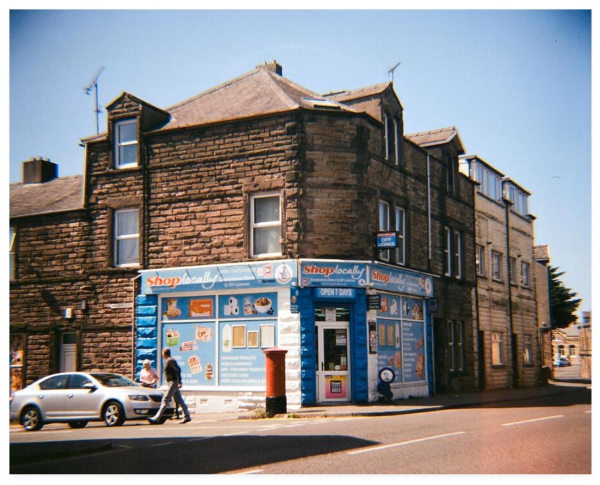 Colour photograph showing a roadside corner shop in Workington, northern England.  The Shop Locally store has advertisements plastered all over its windows (how do they get any natural light in there?) and an 'off licence' sign affixed to the wall.  Outside the door is a red postbox.  A car is parked to the side and a couple are returning to it.  