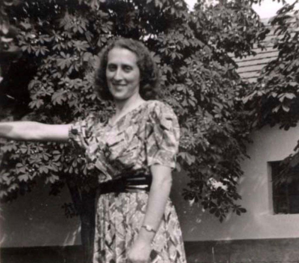 Vintage black and white photo of a woman smiling and standing under a tree, wearing a patterned dress with a belt. A house is partially visible in the background.