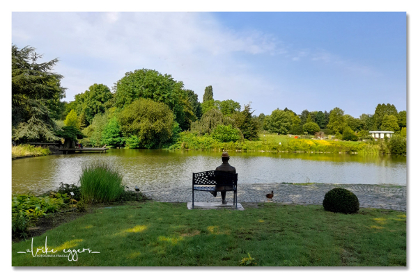 Ein kleiner See, umgeben von Grün (Pflanzen, Büsche, Bäume in verschiedenen Höhen und Farbabstufungen). Am vorderen Ufer steht eine Bank aus Metall mit einer verzierten Lehne, darauf die Bronzeplastik eines Mannes, der sehr entspannt mit übergeschlagenen Beinen auf der Bank sitzt. Jemand hat der Figur einen echten Strohhut auf den Kopf gesetzt. Rechts neben der Bank steht eine Ente. Über allem ein heller Sommerhimmel, zum Teil mit weißen Schleierwolken.