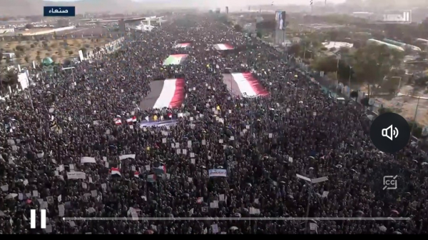 Screenshot from clip: An aerial view of a massive crowd gathered in a public space, stretching far into the distance. Large flags, including Yemeni flags and Palestinian flags, are displayed prominently among the crowd. Many participants hold placards and banners, creating a dense sea of people. The scene takes place in a city with some greenery and urban structures visible in the background. Arabic text appears at the top left corner, and a sound icon is displayed on the right.