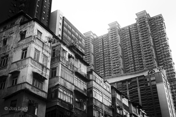 A Grungy Dystopian Black And White Cityscape Photo In Landscape Format. In The Foreground And Stretching From The Upper Left Margin And Descending Towards The Lower Right Corner Is A Row Of Low-Rise Mixed Use (Residential, Business Etc) Buildings Of Perhaps Six To Ten Floors. They All Have A Disheveled And Neglected Appearance. Many Have Randomly Placed External Air Conditioning Units, Clothes Racks And The Occasional Open Window. Just In View In The Upper Left Corner Is A Penthouse Shack, An Addition Made By Slum Landlords For Low Rent Tenants. 
Contrasting In The Background And Predominantly In The Upper Right Corner Are Modern Very High-Rise Apartment Blocks, All Very Uniform And Clean.
The Location Is To Kwa Wan In Kowloon City, Hong Kong. The Low-Rise Buildings Probably Being A Relic From The Former Kai Tai Airport That Was Situated A Few Hundred Metres Away And Would Have Stunted The Vertical Growth Of Any Nearby Buildings (2017).