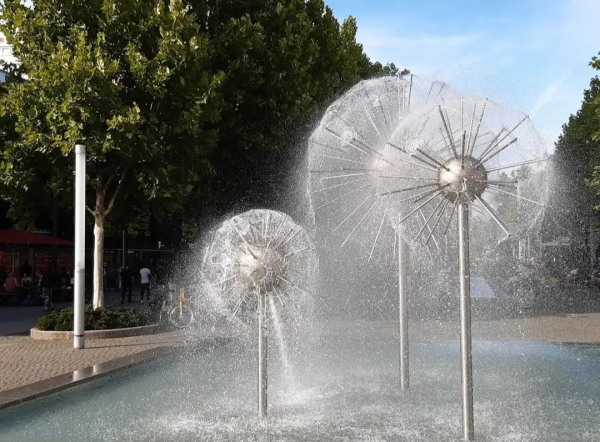 Blick auf einen Brunnen. Aus Kugeln stehen dünne Stäbe hervor, die einen feinen Sprühnebel verteilen. Im Hintergrund Bäume