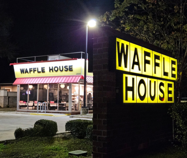 Late night in front of a Waffle House restaurant. Beneath the deep dark night, the iconic building's colors and design glows like a painting under spotlights, just slightly to the right, and in the foreground, the large Waffle House sign, with the iconic yellow scrabble-like squares, each with a letter in black spelling out Waffle House.