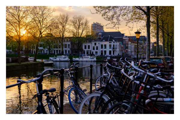 Ein Kanal in Utrecht zur goldenen Stunde. Direkt vor der Kamera stehen Unmengen von Fahrrädern, unter anderem auch am Geländer des Kanals. Im Kanals selbst ist eine Kreuzung, einige Boote sind am Rand angelegt. Auf der anderen Seite des Kanals sind Häuser, über diesen geht die Sonne unter und scheint dabei durch blätterlose Bäume. Dadurch ist alles in goldenes Licht getaucht. 