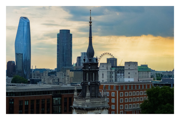 Blick über die Dächer Londons. Direkt vorne mittig ist die Spitze eines alten Kirchturms. Dahinter zeigen sich die Unterschiede Londons, alte Gebäude, neue Wolkenkratzer, heruntergekommene Wohngebäude, des Riesenrad London Eye ist am Horizont. Der ist teils grau, teils tritt das Sonnenlicht durch die Wolken und färbt den Himmel goldgelb. 