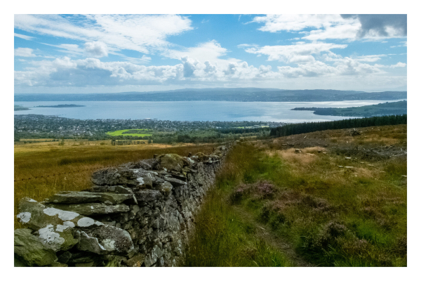 Blick von einem Berg auf eine Stadt am Wasser. Der Berg ist bedeckt mit gelb-grünem Gras, von der linken unteren Ecke aus, geht eine Steinmauer Richtung Tal. Unten im Tal ist eine Stadt an einem See, der übers gesamte Bild geht. Der Himmel ist leicht bewölkt. 