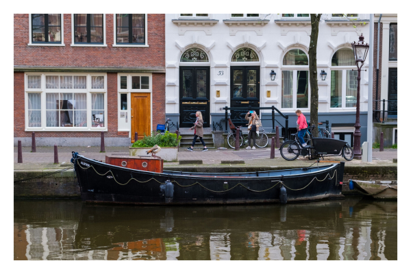 Ein Kanal in Amsterdam. Am Ufer liegt ein offenes Boot mit einem kleinen rotbraunen Häuschen. Darauf sitzt eine Ente. Am Ufer dahinter laufen mehrere Personen, am Straßenrand stehen Fahrräder. Dahinter sind Gebäude. 