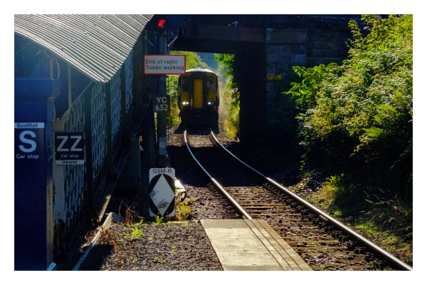 Gleise, ein gelber Zug fährt durch einen kleinen Tunnel in den Bahnhof ein. Rechts ist Gebüsch, links Teil des Bahnhofs. Die Schienen enden genau rechts unten im Bild. 