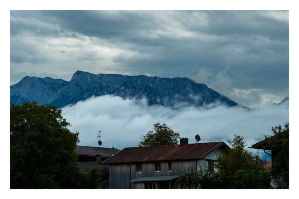 Foto im Querformat. Vorne sind ein paar Häuser, am Horizont die Alpen, dazwischen ist ein Wolkenmeer. Der Himmel über den Alpen ist auch komplett bewölkt. 