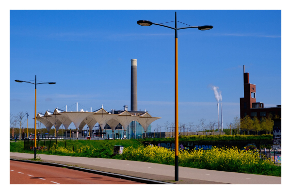 Foto im Querformat. Ein Gewerbe bzw Industriegebiet in Utrecht. Vorne verläuft ein breiter Fahrradweg mit hohen gelben Straßenlaternen, dahinter ist Wiese mit gelben Blumen, dann eine Bahn bzw Busstation mit einem modernen Dach auf mehreren Säulen, dahinter sind ein Industriegäude und mehrere Schornsteine, aus denen teils Rauch austritt. Der Himmel ist strahlend blau. 