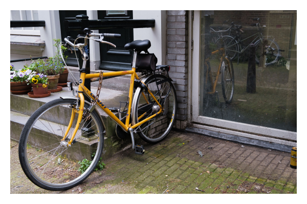 Foto im Querformat. Ein gelbes Fahrrad steht am Geländer eines Treppenaufgangs. Es ist mit einer Kette angeschlossen. Dahinter ist ein Fenster in der Hauswand auf Bodenhöhe. Darin spiegelt sich das Fahrrad. 